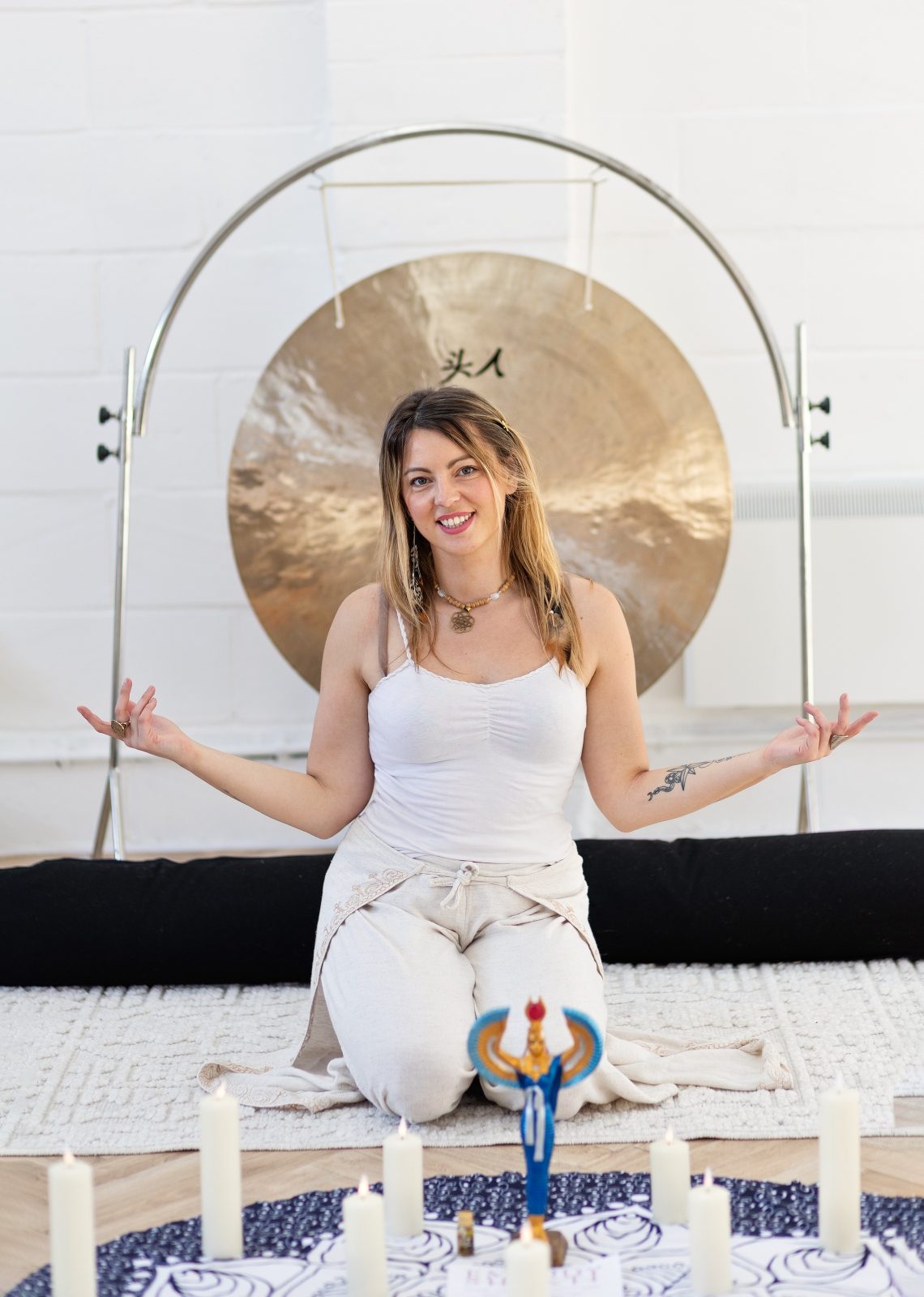 Monika seated before a gong, hands open, candles in the foreground
